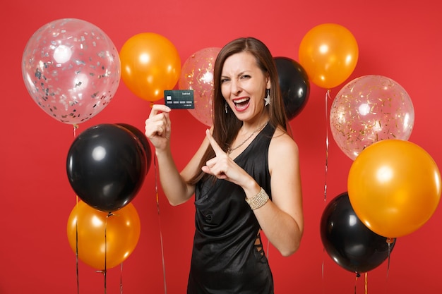 Fröhliches Mädchen im schwarzen Kleid feiert, Zeigefinger auf Kreditkarte in der Hand auf Luftballons mit rotem Hintergrund zeigend. Internationaler Frauentag, Frohes neues Jahr, Geburtstagsmodell-Feiertagsparty-Konzept.