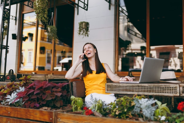 Fröhliches Mädchen im Straßencafé im Freien, das mit Laptop-PC am Tisch sitzt, mit dem Handy spricht, angenehme Gespräche führt, im Restaurant in der Freizeit