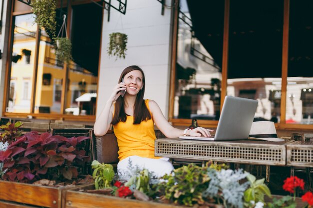 Fröhliches Mädchen im Straßencafé im Freien, das mit Laptop-PC am Tisch sitzt, mit dem Handy spricht, angenehme Gespräche führt, im Restaurant in der Freizeit