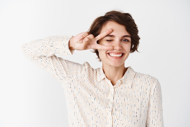 Fröhliches Mädchen in Bluse, das Friedens- oder Victory-Zeichen auf dem Auge zeigt und zwinkert, Positivität und Freude ausdrückt und gegen weiße Wand steht