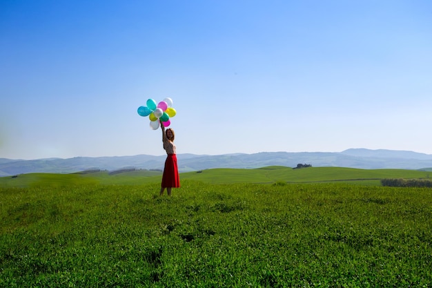 Fröhliches Mädchen in den toskanischen Wiesen mit bunten Luftballons, gegen den blauen Himmel und die grüne Wiese. Toskana, Italien
