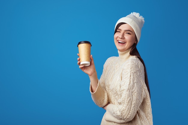 Fröhliches Mädchen in weißem Hut und weißem, warmem Pullover hält eine Tasse zum Mitnehmen