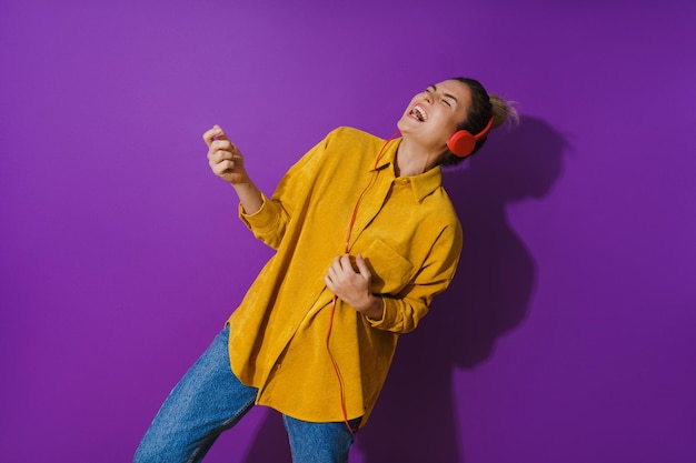 Fröhliches Mädchen mit gelbem Hemd, das Musik mit roten Kopfhörern hört und Luftgitarre vor violettem Hintergrund spielt