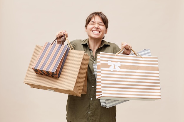 Fröhliches Mädchen mit geschlossenen Augen, das sich über das Einkaufen freut und abgestreifte Taschen gegen die beige Wand hält