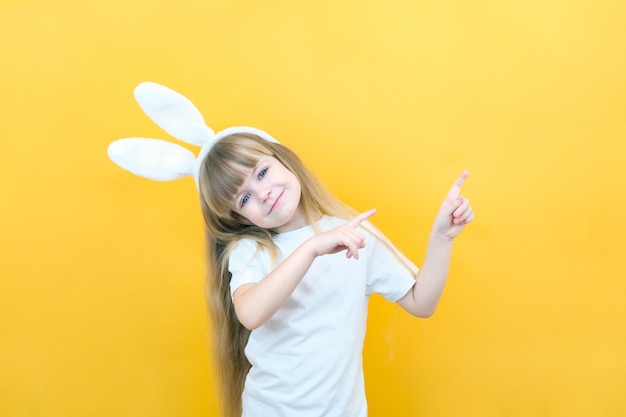 Fröhliches Mädchen mit Hasenohren auf dem Kopf auf gelbem Hintergrund Lustiges glückliches Kind zeigt mit den Fingern auf einen leeren Raum, kopieren Sie Platz für Text-Mockup