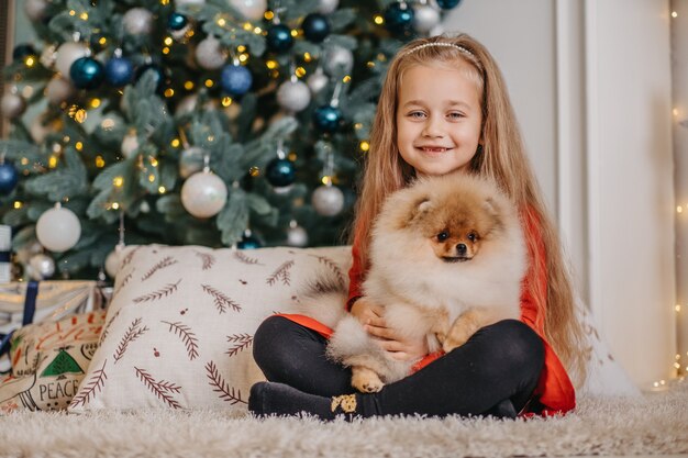 Fröhliches Mädchen posiert mit süßem Welpen, Weihnachtsbaum im Hintergrund, lang erwartetes flauschiges Geschenk, Freude