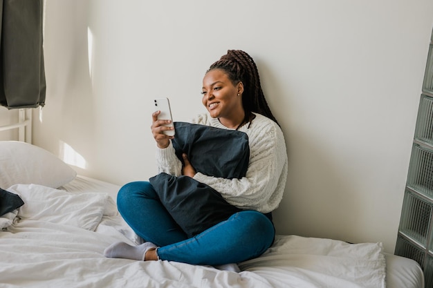 Fröhliches modernes afroamerikanisches junges Mädchen, das über Video-Chat spricht und zu Hause sitzt Kommunikationskonzept Lächelndes Hipster-Mädchen, das sich zu Hause auf ihrem Telefon selbst porträtiert
