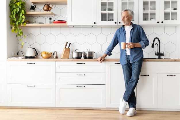 Fröhliches Morgenporträt eines lächelnden älteren Mannes, der in der Küche Kaffee trinkt