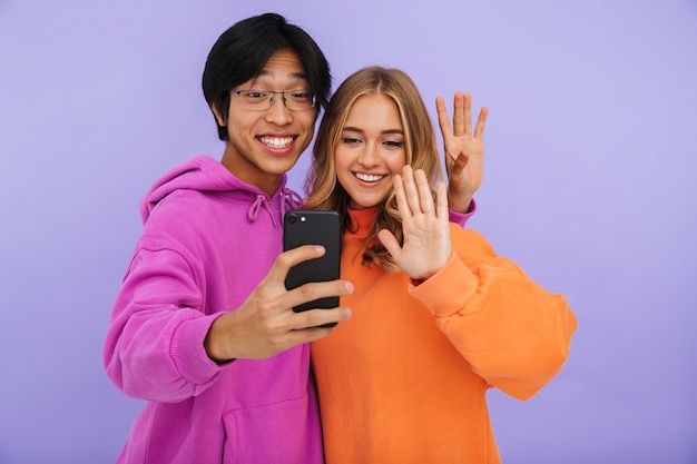 Fröhliches multhiethnisches Teenagerpaar, das isoliert zusammen steht und ein Selfie macht