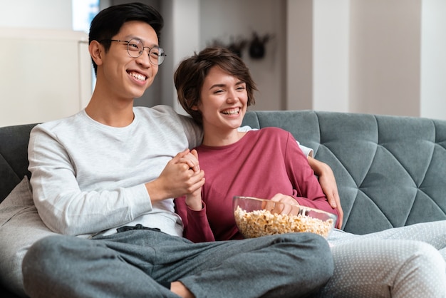 Fröhliches multinationales Paar, das Popcorn isst und einen Film schaut, während es zu Hause auf dem Sofa sitzt