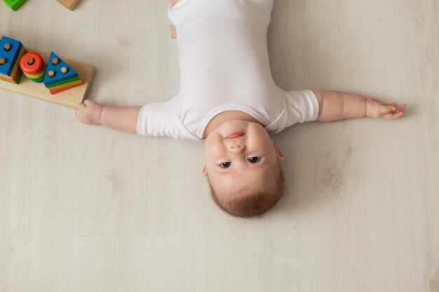 Fröhliches Neugeborenes in einem weißen Bodysuit liegt auf dem Rücken auf dem Boden und spielt mit Lernspielzeug. Produkte für Kinder. konzept einer glücklichen kindheit und mutterschaft. Kinderbetreuung. Platz für Text