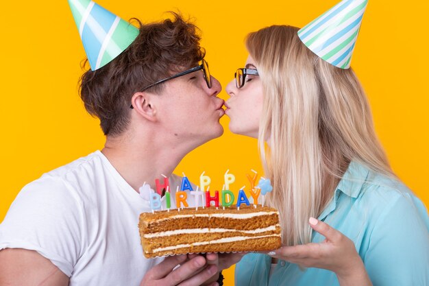 Fröhliches Paar charmanter Kerl und süßes Mädchen in Papierhüten machen ein dummes Gesicht und halten in ihren Händen Kuchen mit der Aufschrift Geburtstag auf gelbem Hintergrund. Konzeptgrüße und Streich.