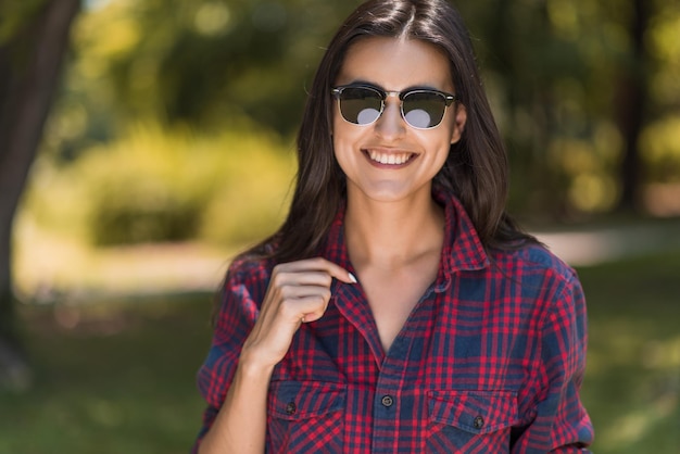 Fröhliches Porträt einer jungen, brünetten, kaukasischen Frau, die vor Naturhintergrund im Park lächelt und eine Sonnenbrille trägt Lifestyle-Konzept
