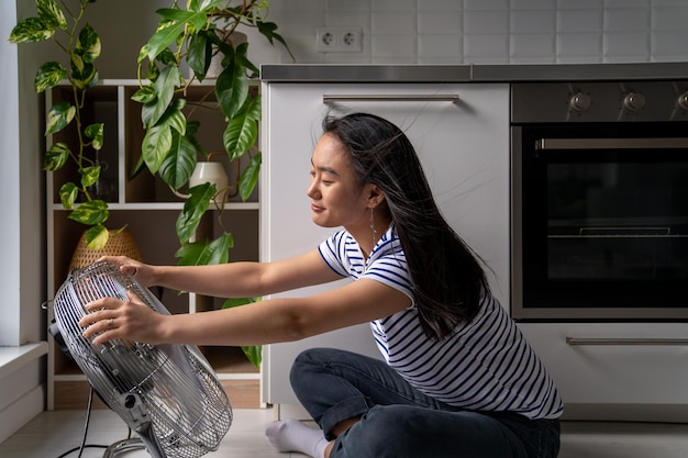 Fröhliches, positives asiatisches Mädchen genießt den kalten Wind vom elektrischen Ventilator, der auf dem Boden in der Küche des Hauses sitzt