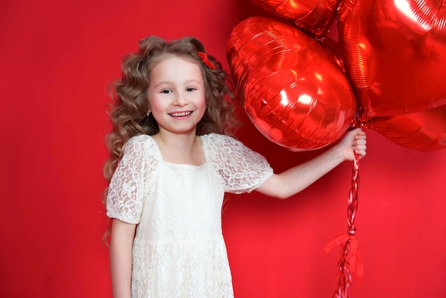 Fröhliches positives, fröhliches, kleines Mädchen im Kleid, das rote herzförmige Luftballons hält