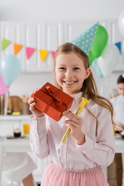 fröhliches Preteen-Mädchen mit Geburtstagsgeschenk und Party-Horn zu Hause
