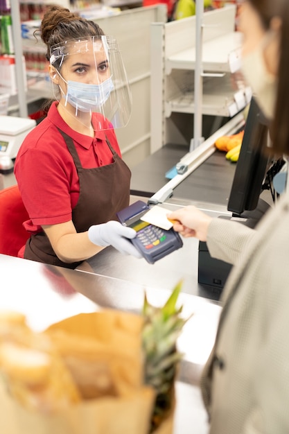 Fröhliches reifes Paar, das an der Registrierkasse vor einer jungen Kassiererin mit gemischten Rassen steht und scannt, was sie im Supermarkt gekauft haben