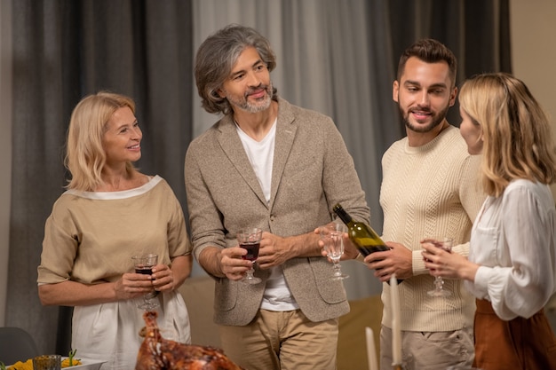 Fröhliches reifes Paar mit Gläsern Rotwein und junger Mann mit einer Flasche Alkohol mit Blick auf die blonde Frau, die Toast am servierten festlichen Tisch macht