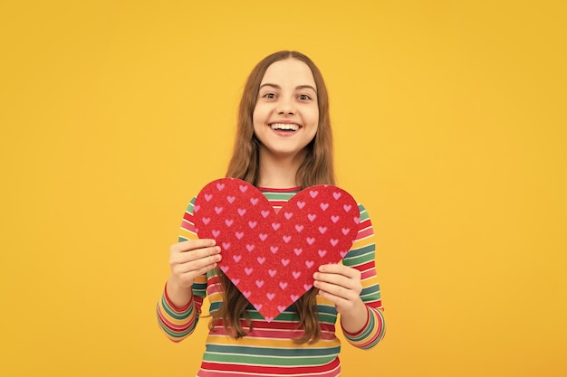 Fröhliches, reizendes, romantisches Teenager-Mädchen hält rotes Herzsymbol der Liebe zum Valentinstag isoliert auf gelbem Hintergrund