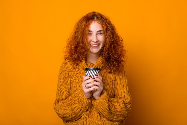 Fröhliches rötliches Mädchen, das in die Kamera lächelt und eine Tasse Kaffee hält, isolierter Hintergrund des Überstands