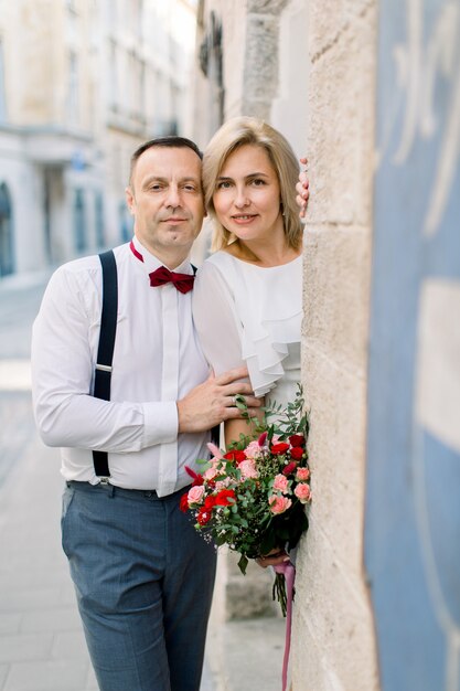 Fröhliches romantisches Paar mittleren Alters, gutaussehender Mann in eleganter Kleidung und hübsche Dame in weißem Kleid und Blumenstrauß, posiert mit Blick auf die Kamera in der Nähe des alten Stadtgebäudes