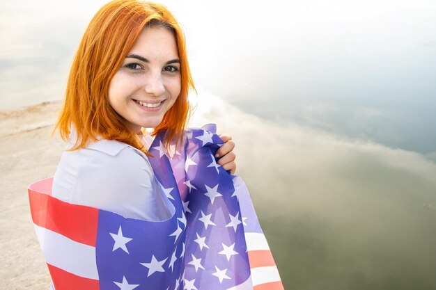 Fröhliches rothaariges Mädchen mit US-Nationalflagge auf den Schultern Positive junge Frau, die den Unabhängigkeitstag der Vereinigten Staaten feiert Internationaler Tag der Demokratie Konzept