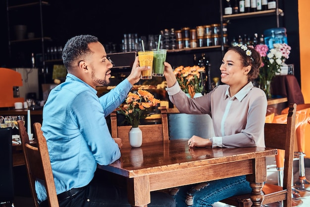 Fröhliches schönes afroamerikanisches verliebtes Paar, das eine tolle Zeit zusammen in einem Restaurant bei ihrer Verabredung hat, ein attraktives Paar, das sich amüsiert, hält Gläser Saft.