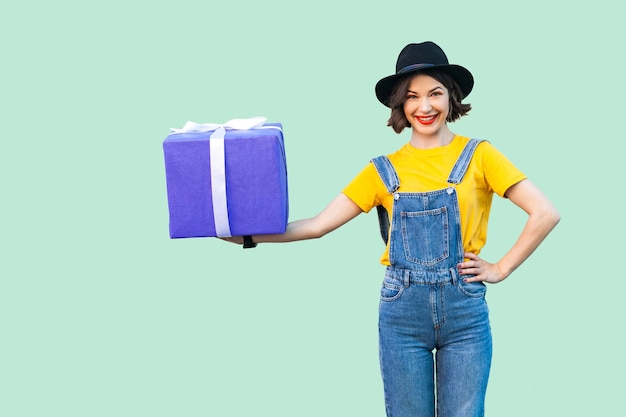 Fröhliches, schönes junges mädchen in hipster-kleidung in denim-overalls und schwarzem hut, das eine große schwere geschenkbox mit einem zahnigen lächeln mit der hand am gürtel hält und in die kamera schaut. studioaufnahme, grüner hintergrund