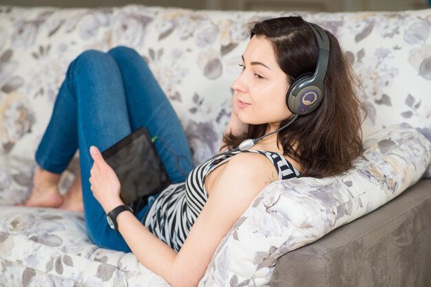 Fröhliches, schönes junges Mädchen mit Kopfhörern genießt zu Hause ein Tablet auf der Couch
