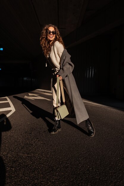 Fröhliches, schönes, lockiges Mädchen mit einem Lächeln mit Sonnenbrille in einem modischen langen Mantel mit einer stilvollen Handtasche in der Stadt auf Sonnenlicht und Schatten. Lustige Frau im lässigen urbanen Stil im Freien