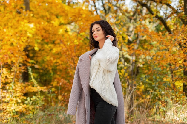 Fröhliches, schönes, lockiges Mädchenmodell in modischer Herbstkleidung mit einem weißen Vintage-Pullover und stilvollem Mantel geht in einem goldenen Herbstpark spazieren