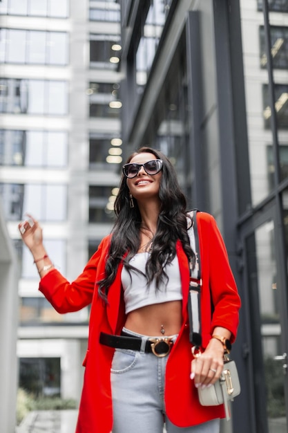Fröhliches, schönes, modisches Geschäftsfrauenmodell mit einem Lächeln und Sonnenbrille in Mode, lässige, stilvolle Kleidung mit rotem Blazer-Oberteil und Jeans mit Tasche geht in der Stadt auf urbanem Hintergrund spazieren