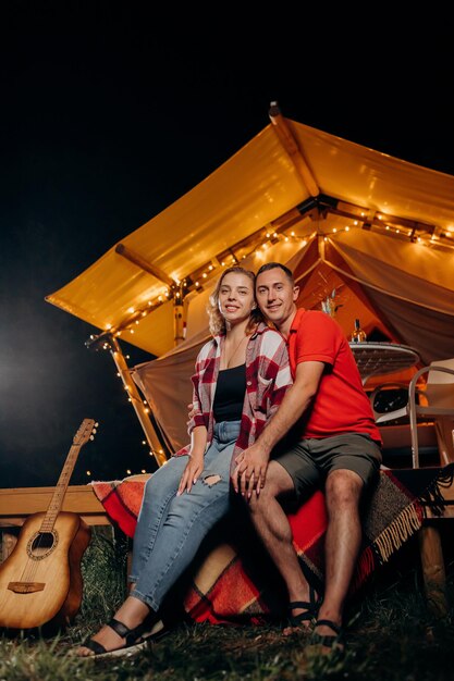 Fröhliches schönes Paar, das sich am Abend in der Nähe des gemütlichen Lagerfeuers im Glamping entspannt Luxus-Campingzelt für Erholung im Freien und Erholung Lifestyle-Konzept