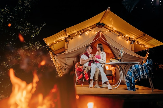 Fröhliches schönes Paar, das sich am Sommerabend im Glamping entspannt und Wein in der Nähe des gemütlichen Lagerfeuers trinkt Luxus-Campingzelt für Erholung im Freien und Erholung Lifestyle-Konzept