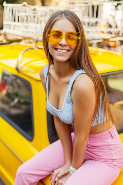 Fröhliches schönes süßes Mädchen mit einem natürlichen Lächeln mit cooler modischer gelber Brille in einem modischen Strickoberteil und stylischen rosa Jeans sitzt auf einem gelben Retro-Auto und genießt