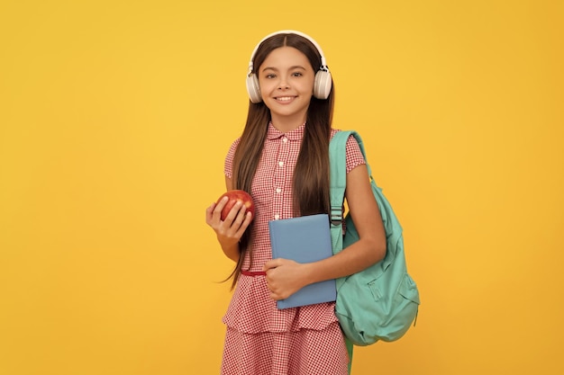 Fröhliches Schulkind mit Kopfhörern trägt Rucksack und Arbeitsbuch mit Apfel-Mittagessen