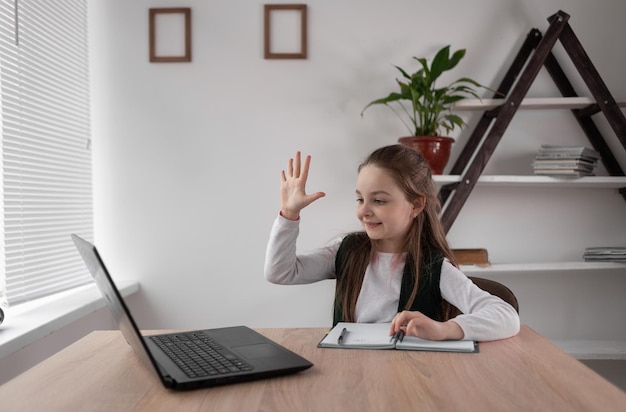 Fröhliches Schulmädchen auf Videokommunikation Online-Unterricht von einem Laptop zu Hause sitzend an einem Schreibtisch mit einem Notizbuch und einem Stift Lehrer aus der Ferne Fernunterrichtskonzept