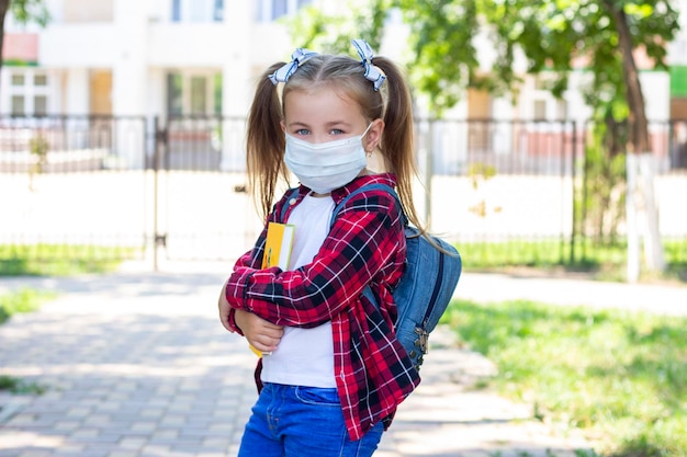 Fröhliches Schulmädchen in Schutzmaske mit Rucksack und Lehrbuch in den Händen. in einem weißen T-Shirt und einem karierten Hemd