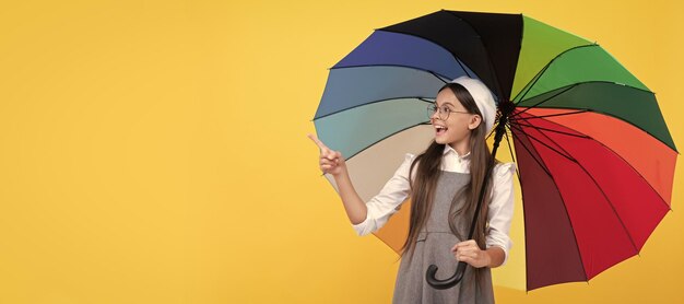 Fröhliches Schulmädchen mit Brille Teenager-Kind unter buntem Sonnenschirm, das auf Finegr zeigt Kind mit Regenschirm Regenwetter horizontales Plakatbanner mit Kopierraum