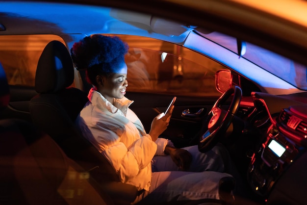 Fröhliches schwarzes Mädchen sitzt im Auto und lädt junge afroamerikanische Smartphone-Süchtige in soziale Medien hoch