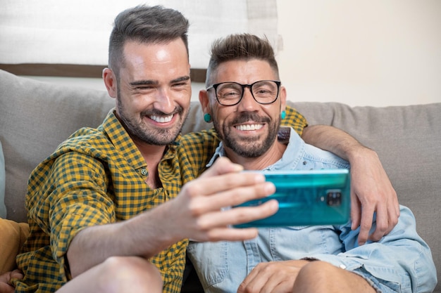 Fröhliches schwules Paar, das auf der Couch sitzt, sich umarmt und auf das Handy schaut und einen Film anschaut, der durch das soziale Netzwerk scrollt