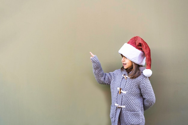 Fröhliches, selbstbewusstes kleines Mädchen mit Weihnachtsmann-Hut, das auf grünen Kopierraum zeigt