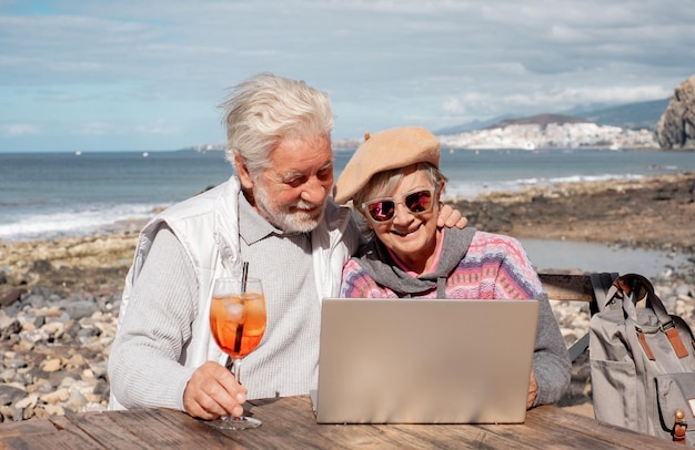 Fröhliches Seniorenpaar, das gemeinsam auf dem Laptop surft und den sonnigen Tag im Freien am Strand genießt