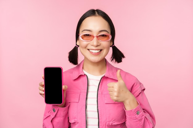 Fröhliches stilvolles Mädchen empfiehlt die Anwendung auf dem Mobiltelefon Lächelndes asiatisches weibliches Modell mit Smartphone-Bildschirm und Daumen nach oben, das über rosafarbenem Hintergrund steht
