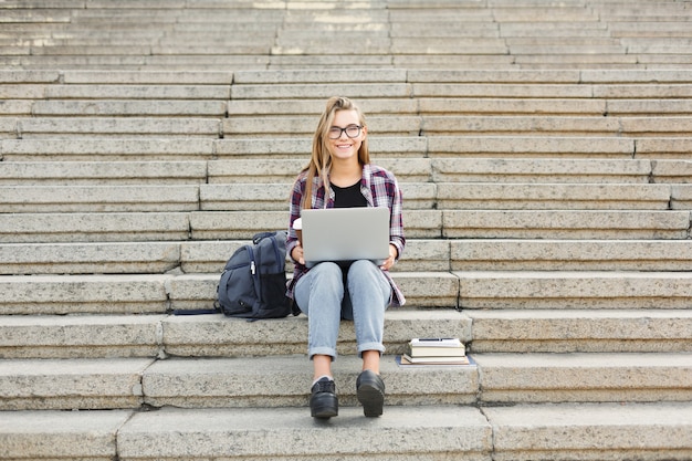 Fröhliches Studentenmädchen, das auf der Treppe sitzt und mit Laptop arbeitet, sich auf Prüfungen vorbereitet und draußen Kaffee trinkt, auf dem Universitätscampus. Technologie-, Bildungs- und Fernarbeitskonzept, Kopierraum