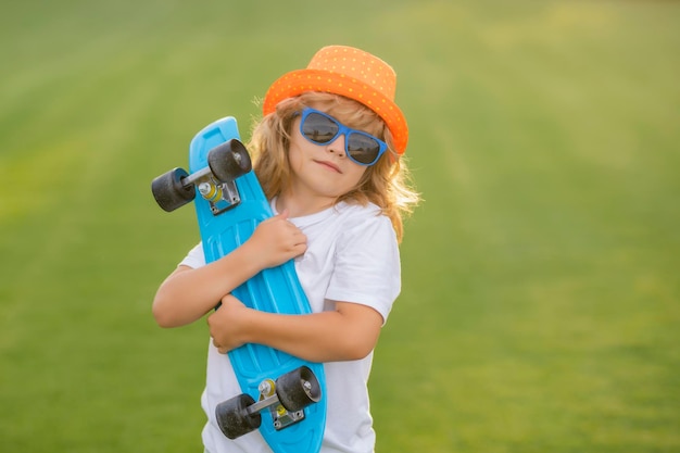 Fröhliches, stylisches Kind mit Sonnenbrille und modischem Sommerhut posiert mit Skateboard im Freien