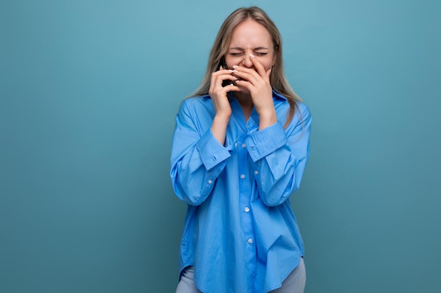 Fröhliches, süßes blondes Mädchen lacht und bedeckt ihren Mund, während sie auf einem blauen, hellen Hintergrund telefoniert