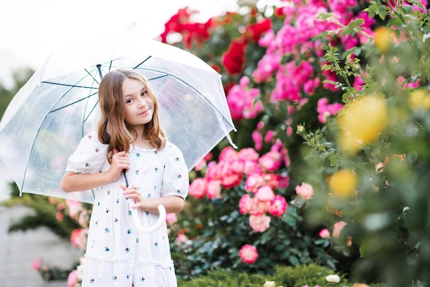 Fröhliches süßes Kindermädchen mit langen blonden Haaren, das einen transparenten Regenschirm hält, der über einer Rosenblume posiert