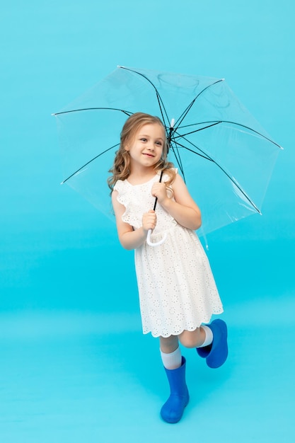 Fröhliches süßes kleines Mädchen in blauen Gummistiefeln und einem baumwollweißen Kleid, das einen Regenschirm auf blauem Hintergrund im Studio hält und lächelt und Platz für Text macht