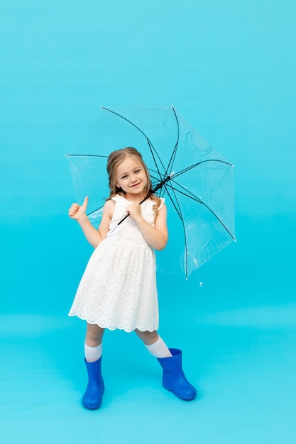 Fröhliches süßes kleines Mädchen in blauen Gummistiefeln und einem baumwollweißen Kleid, das einen Regenschirm auf blauem Hintergrund im Studio hält und lächelt und Platz für Text macht